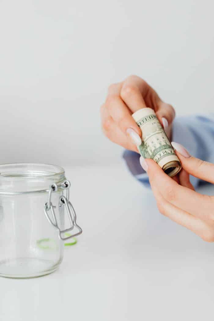 Hands holding and rolling a US dollar bill next to an empty glass jar on a white surface.