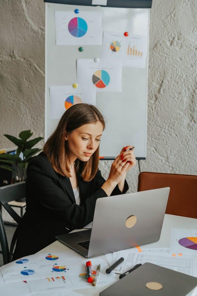 Businesswoman reviewing charts at a laptop in an office setting with presentations behind her.